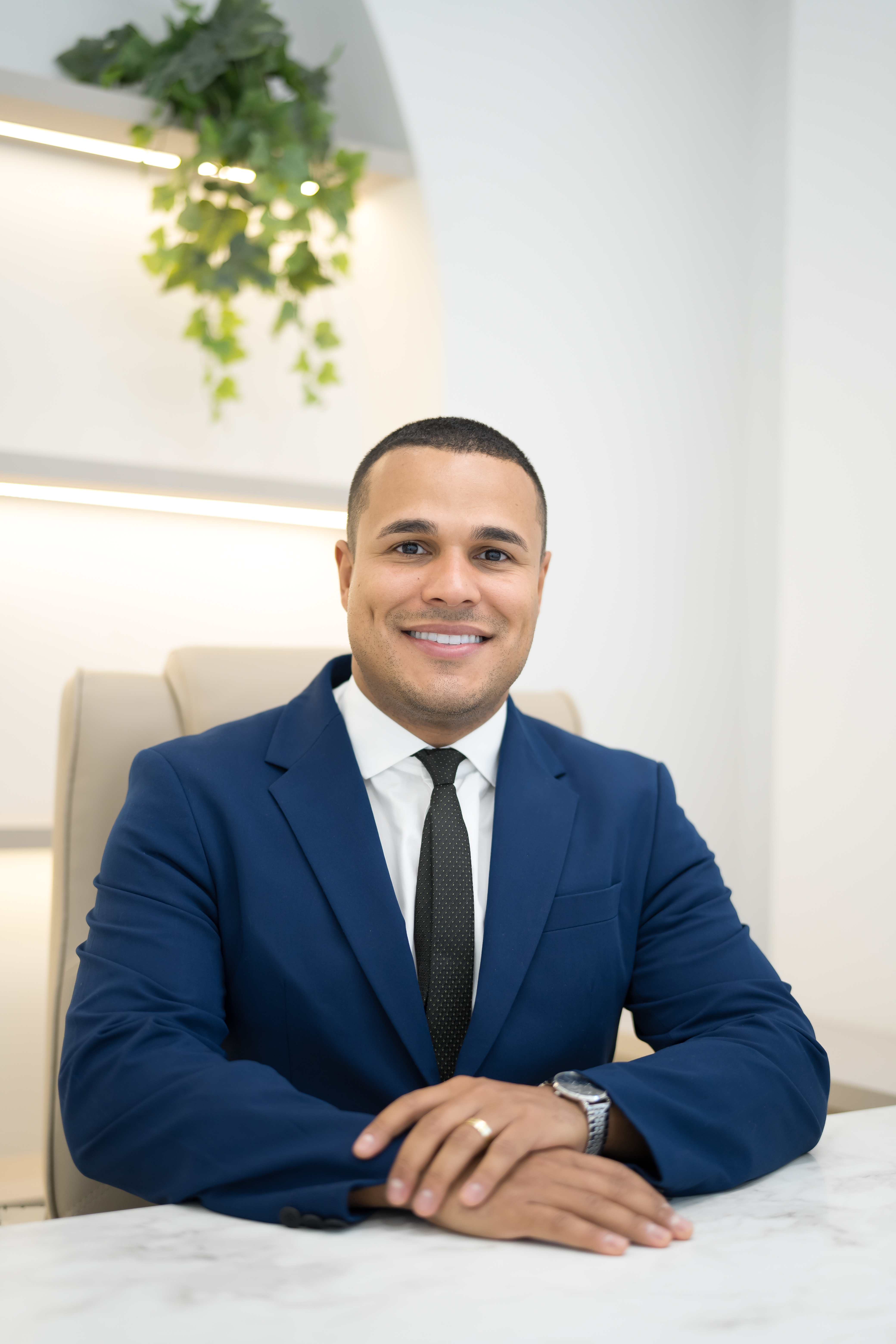 Dr. Marcelo Soares, ortopedista, sorrindo de terno azul-marinho, sentado em cadeira de consultório com planta pendente ao fundo, mãos entrelaçadas sobre mesa de mármore branco, fotografia profissional ultra-realista