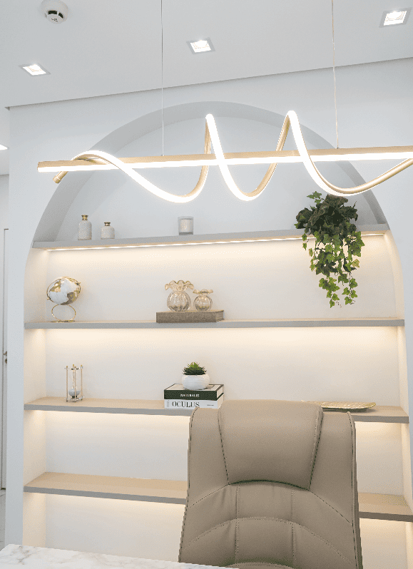 Nicho arqueado de consultório com prateleiras iluminadas por LED embutido, vasos de cerâmica branca, globo terrestre dourado, planta pendente verde e cadeira de couro bege sob luminária ondulada dourada, ambiente clean e sofisticado, fotografia de interiores ultra-realista