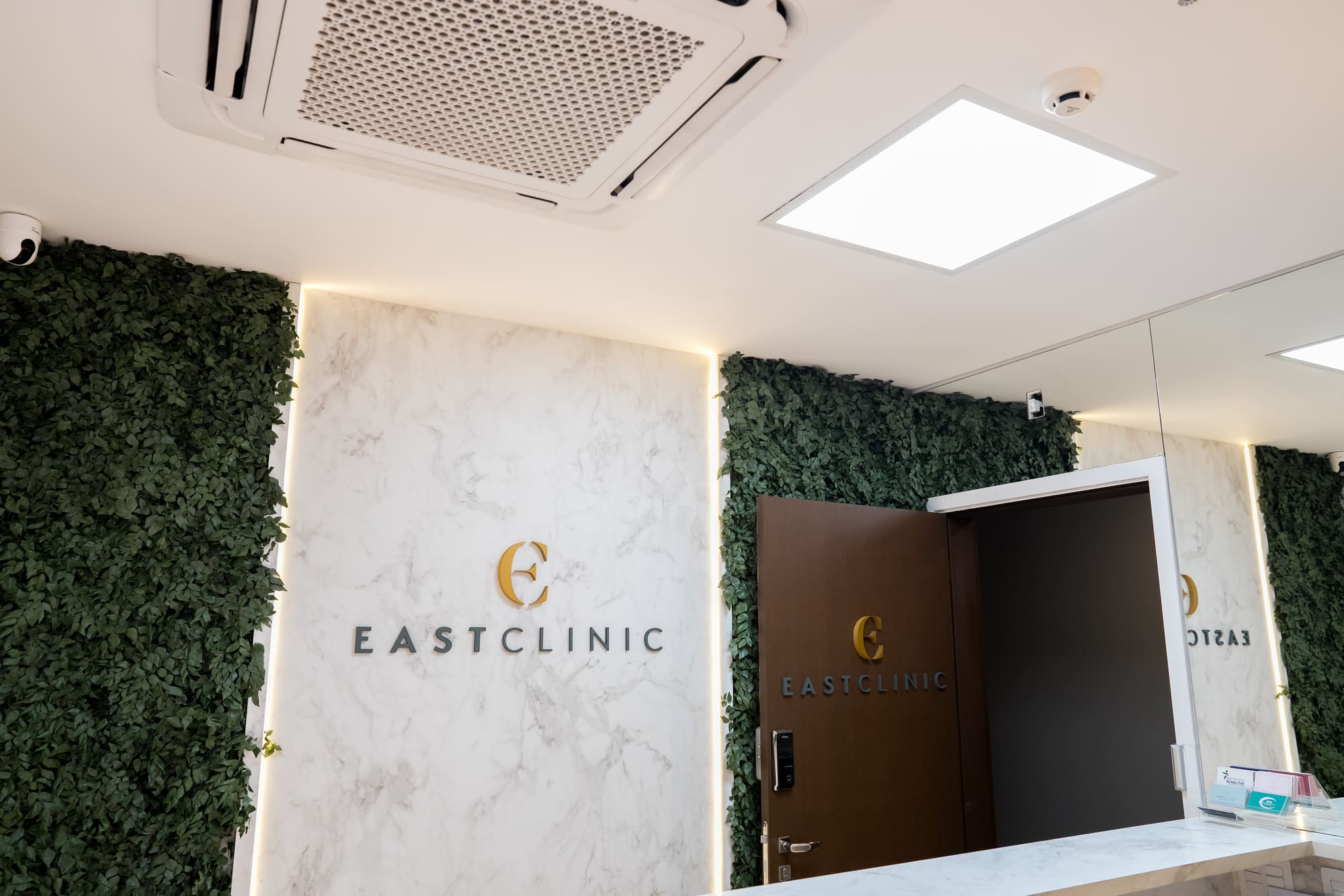 Recepção da EastClinic com parede de mármore iluminada, painel verde de folhagem e logotipo dourado EastClinic, ambiente clean e sofisticado, fotografia de interiores ultra-realista